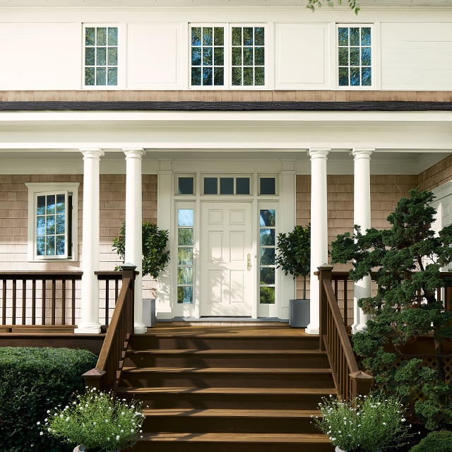 This lovely home features white-painted siding, taupe stained shingles and a brown staircase leading to a front porch with brown railings, white columns and a white front door. This lovely home features white-painted siding, taupe stained shingles and a brown staircase leading to a front porch with brown railings, white columns and a white front door.