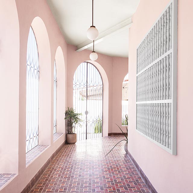 An enclosed porch hallway with pink-painted walls, a white ceiling, arch windows, a light-gray window grating wall piece, and multi-red tile floor. An enclosed porch hallway with pink-painted walls, a white ceiling, arch windows, a light-gray window grating wall piece, and multi-red tile floor.