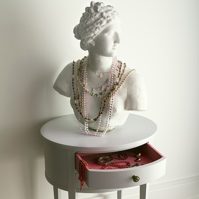 A carved bust with jewelry draped over it on a light gray-painted round table with an open drawer, in front of a white-painted wall. A carved bust with jewelry draped over it on a light gray-painted round table with an open drawer, in front of a white-painted wall.