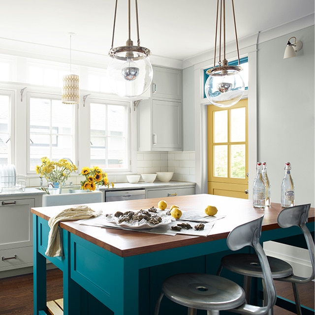 White kitchen with blue-painted kitchen island, yellow door, modern light fixtures, and large windows. White kitchen with blue-painted kitchen island, yellow door, modern light fixtures, and large windows.