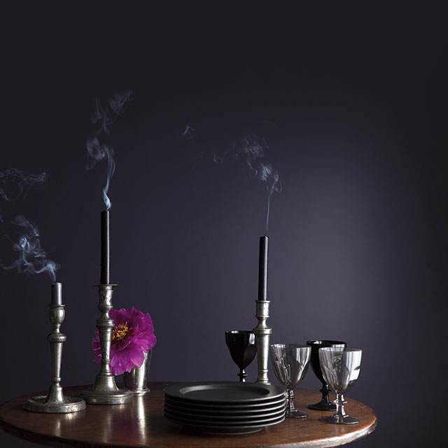 An oval wooden table with just blown-out candles, small plates, and silver goblets against a deep purple wall. An oval wooden table with just blown-out candles, small plates, and silver goblets against a deep purple wall.