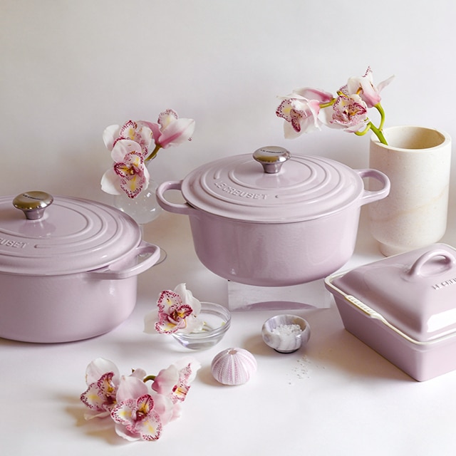 Un éventail de plats de cuisson Le Creuset de couleur Shallot - un charmant gris lavande - agrémenté de fleurs fraîchement coupées se découpe sur un fond blanc cassé. Un éventail de plats de cuisson Le Creuset de couleur Shallot - un charmant gris lavande - agrémenté de fleurs fraîchement coupées se découpe sur un fond blanc cassé.