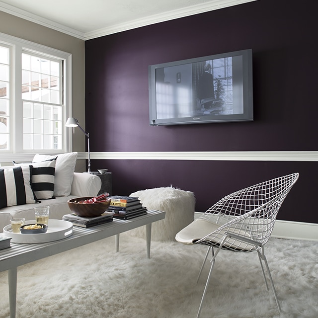 A contemporary living room with a dark purple accent wall frames a flat-screen TV, a white couch with black and white throw pillows, white metal chair, shaggy white rug, and two faux fur ottomans. A contemporary living room with a dark purple accent wall frames a flat-screen TV, a white couch with black and white throw pillows, white metal chair, shaggy white rug, and two faux fur ottomans.